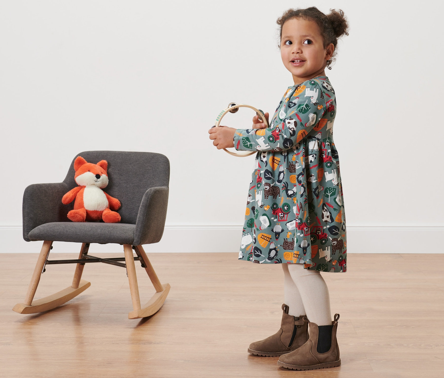 Mädchen im Baby Jerseykleid mit Alloverprint hält ein Tamburin. Daneben steht ein grauer Schaukelstuhl mit einem Plüschfuchs.