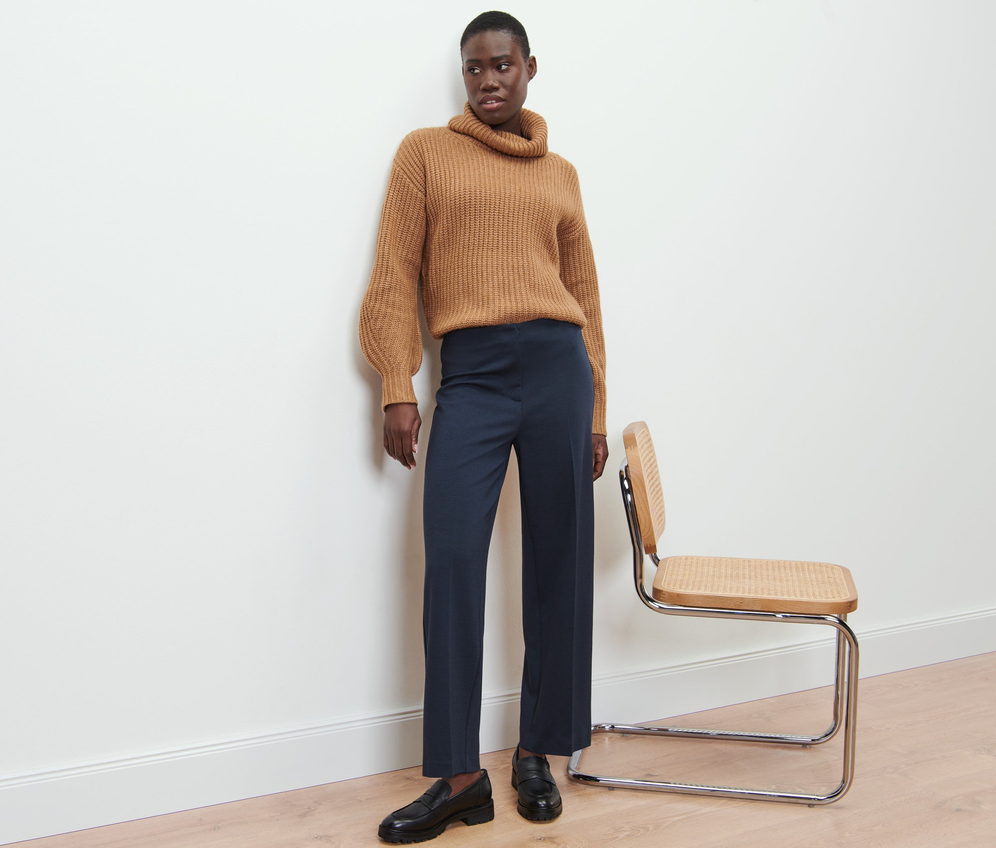 Eine Schwarze steht an einer Wand und trägt einen braunen Rollkragenpullover und eine blaue melierte Jerseyhose.