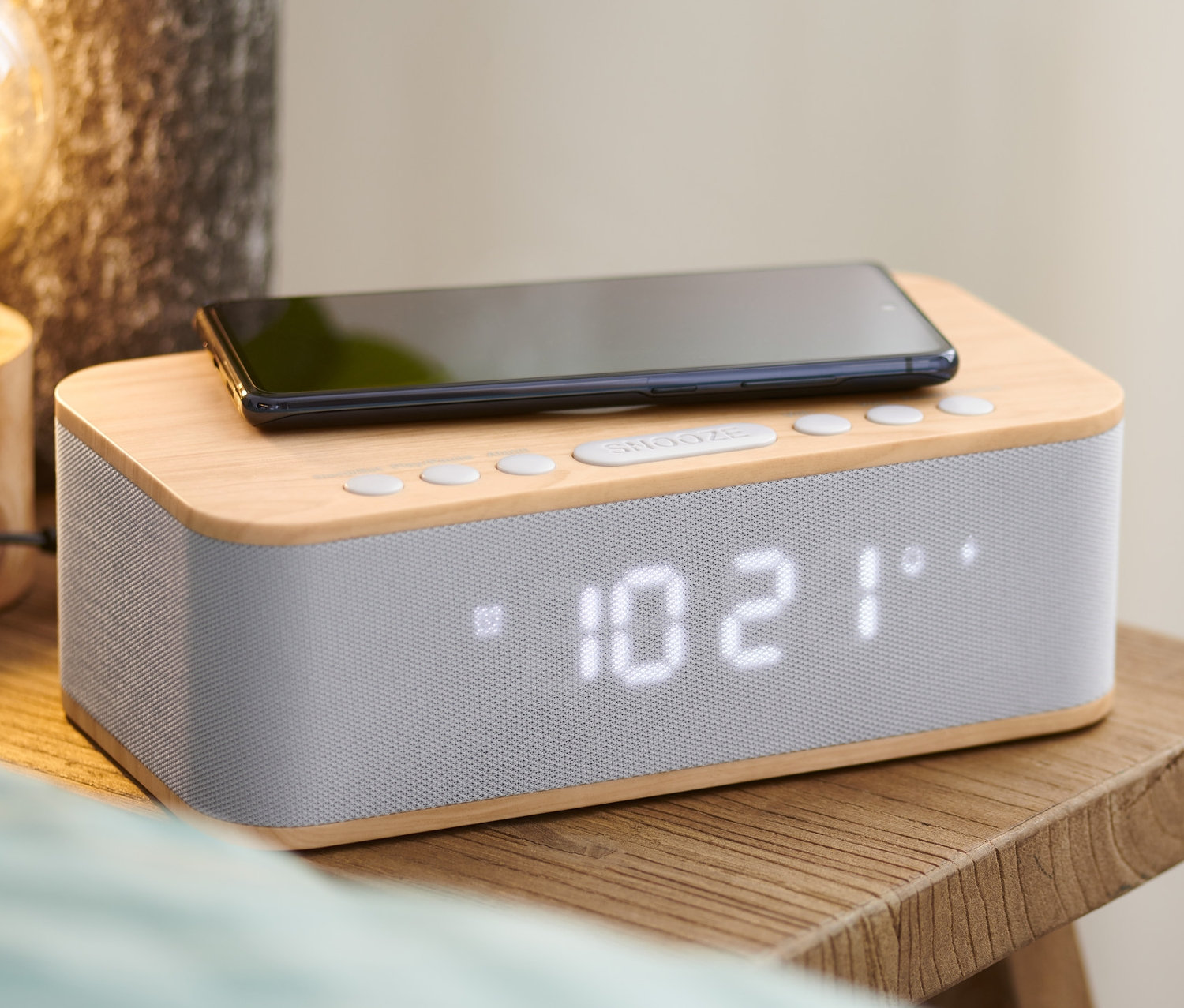 Schwarzes Smartphone liegt auf Wecker mit kabellosem Laden und Bluetooth® auf einem Nachttisch aus Holz.