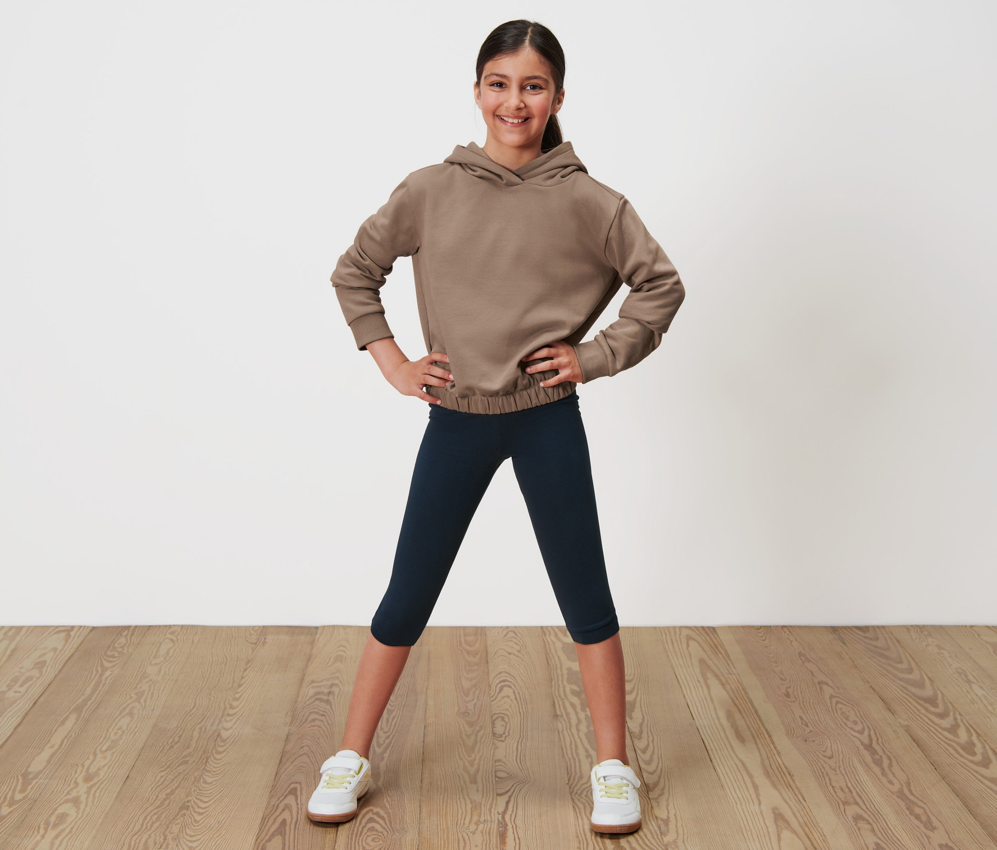 Mädchen posiert in braunem Hoodie und dunkelblauen 3/4-Leggings.