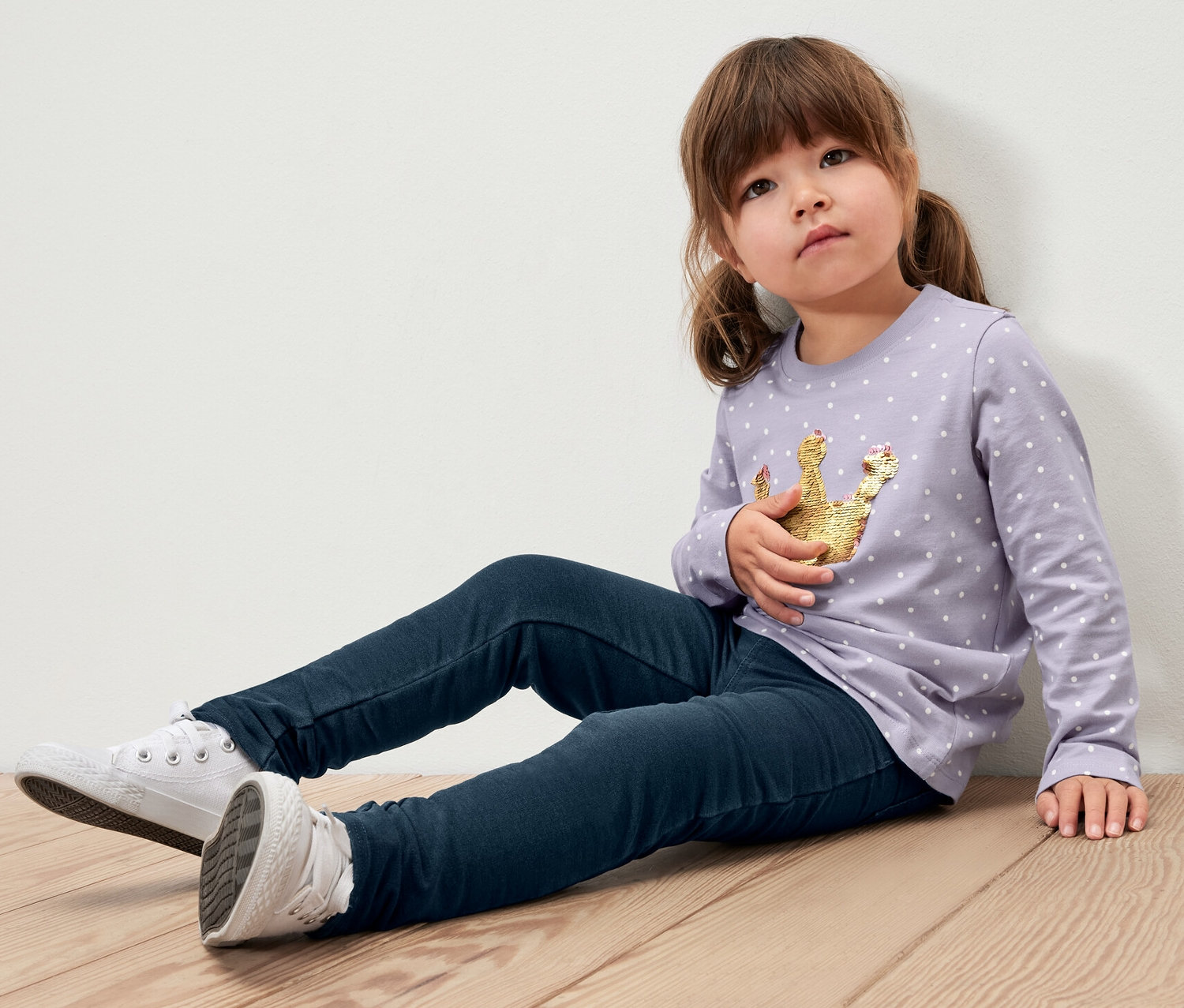 Ein Mädchen sitzt auf einem Holzboden und trägt 2 Kinder-Jeggings ‒ Fit »Mia«, ein lila farbenes Hemd mit einer Krone und weiße Turnschuhe.