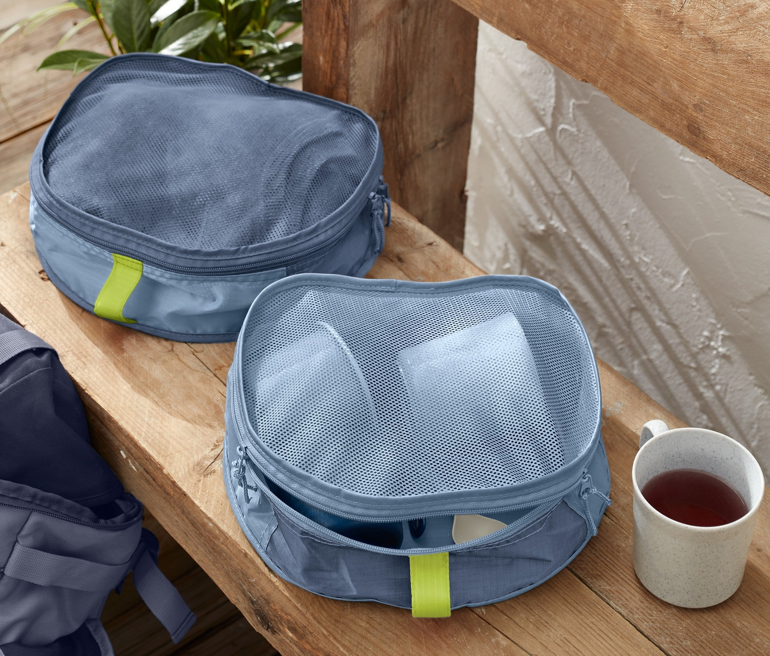 Zwei Rucksack-Organizer liegen auf einer Holzbank neben einer Tasse. Im Hintergrund ist ein Rucksack zu sehen.