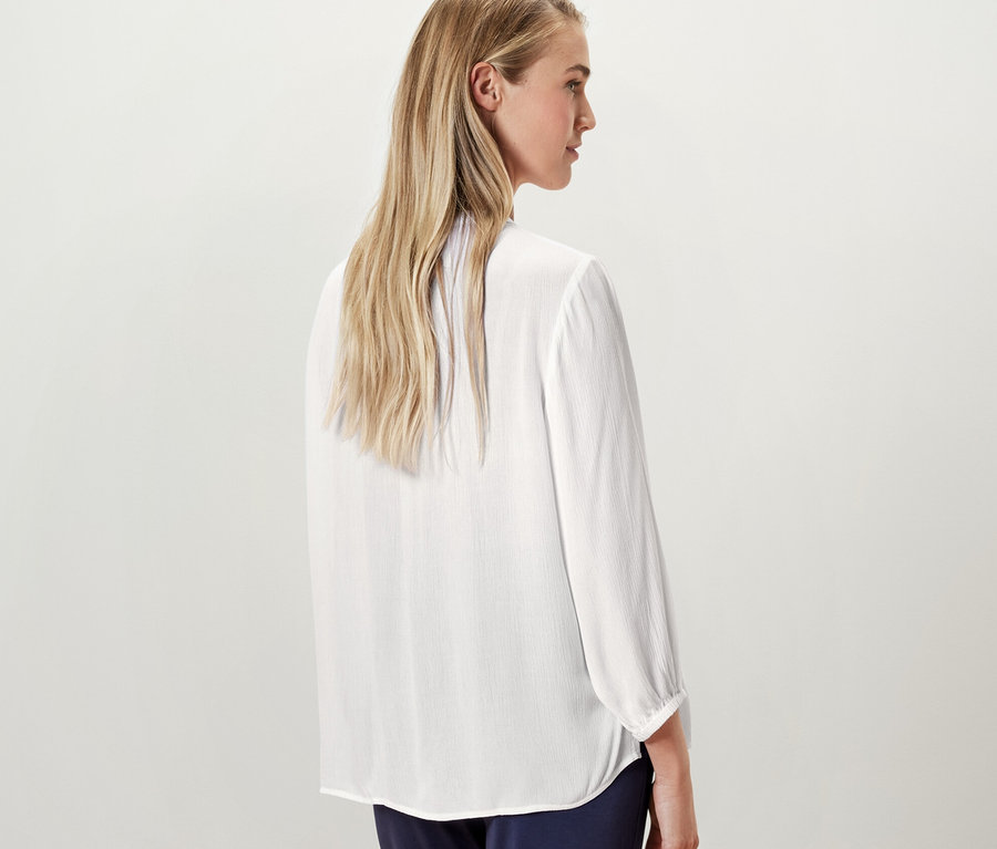 Blonde Frau posiert mit dem Rücken zur Kamera und trägt eine weiße Tunika-Bluse.