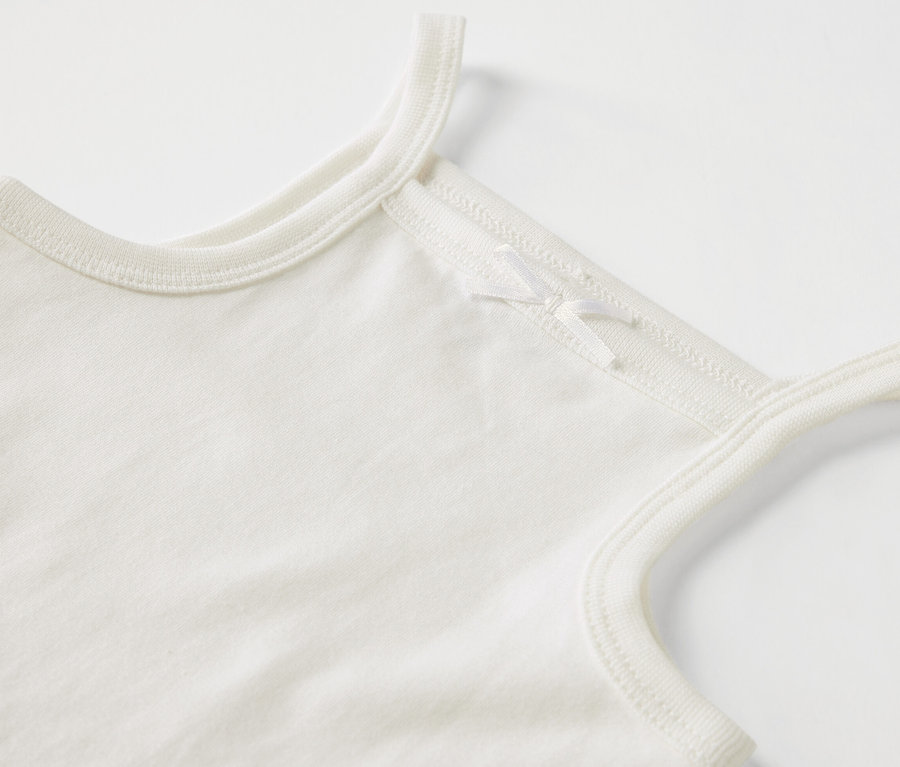 Detailaufnahme eines weißen Baby-Bodies mit Trägern und Schleife.