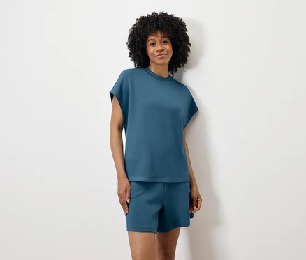 Frau mit lockigem Haar, in blauem T-Shirt und Shorts, steht vor einer weißen Wand.