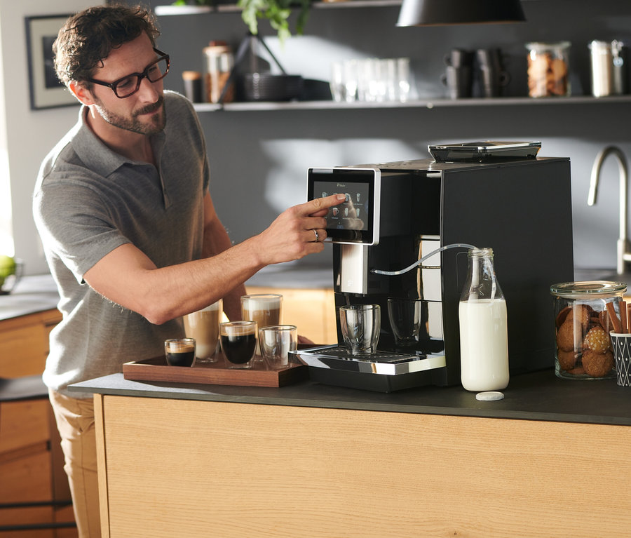 Mann bedient Tchibo Office Kaffeevollautomat, schwarz. Auf dem Tisch stehen Kaffeetassen und Milch.