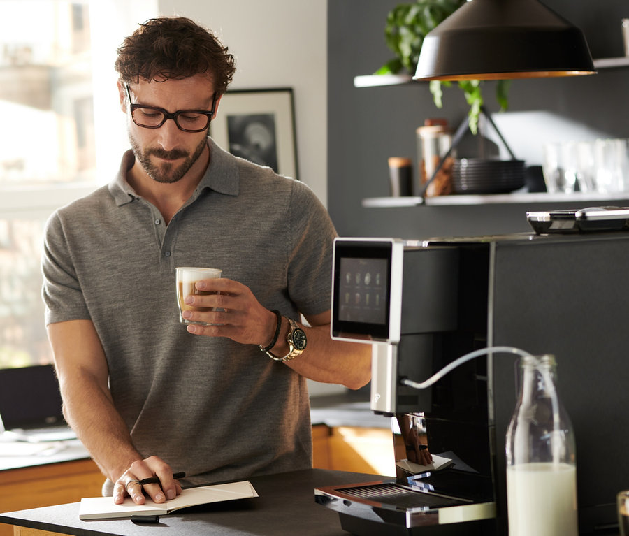 Mann trinkt Kaffee neben »Tchibo Office« Kaffeevollautomat, schwarz.