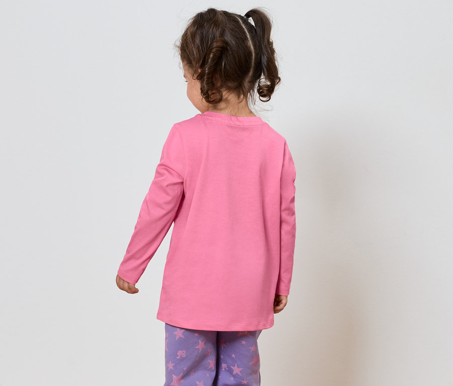 Kleines Mädchen mit braunen Haaren, gekleidet in einem rosa Langarmshirt und einer lila Hose mit Sternenmuster, steht mit dem Rücken zum Betrachter.