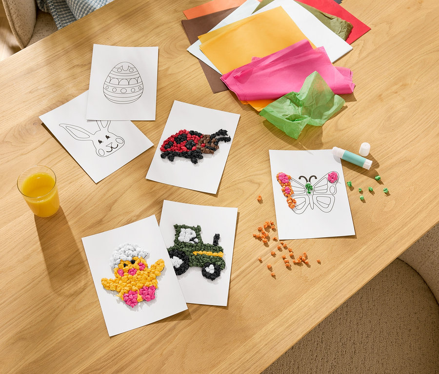 Auf einem Holztisch liegen Bastelarbeiten, darunter eine beklebte Küken-, Traktor-, Schmetterlings- und Schildkrötenkarte. Buntes Seidenpapier, ein Klebestift und ein Glas Saft sind auch zu sehen.