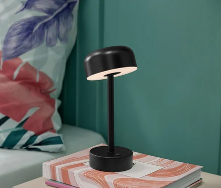 Eine schwarze LED-Akku-Tischleuchte steht auf einem Buch, im Hintergrund ein blumiges Kissen.