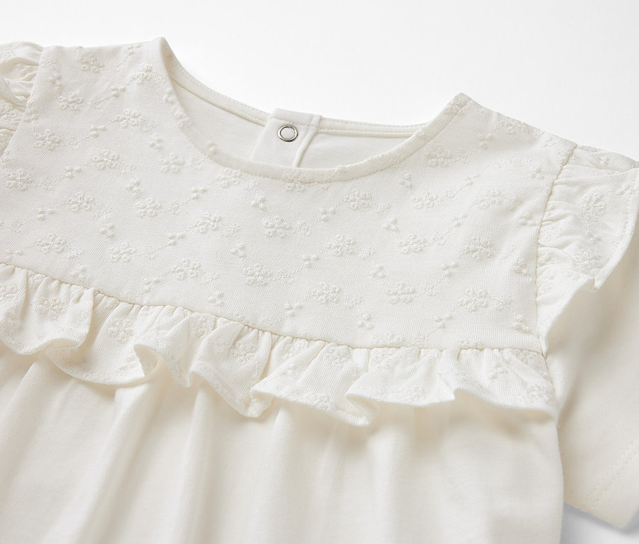 Detailaufnahme eines cremefarbenen Kinder-T-Shirts mit kurzen Ärmeln, Zierrüschen und Stickereien auf der Brust.
