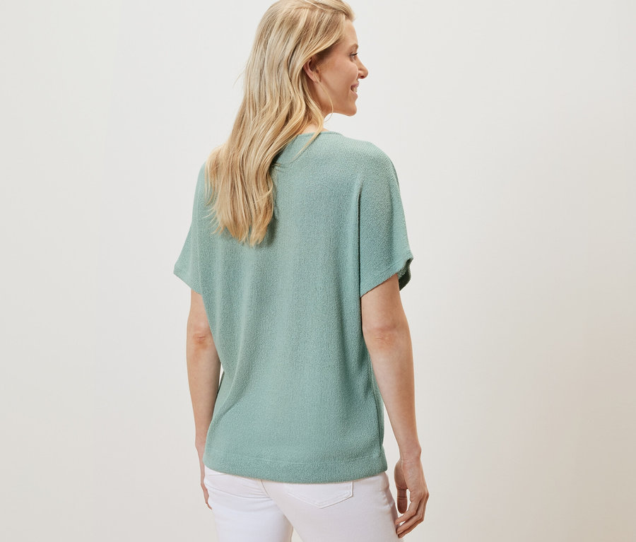 Rückansicht einer blonden Frau, die ein strukturiertes Shirt in Salbei und weiße Hosen trägt.