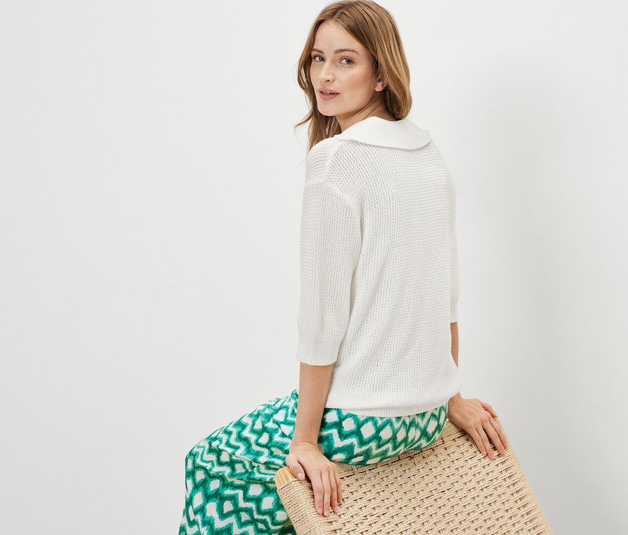 Frau sitzt auf einem Hocker und trägt einen weißen sommerlichen Strickpullover.
