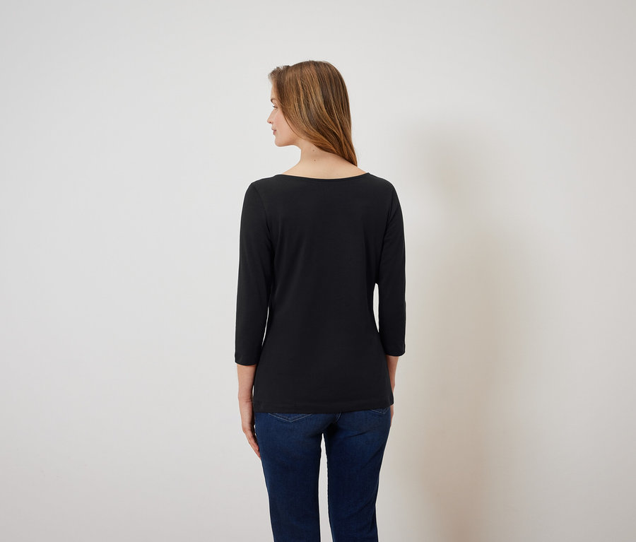 Eine Frau mit braunen Haaren, von hinten gesehen, trägt ein schwarzes Dreiviertelarm-Shirt und blaue Jeans.