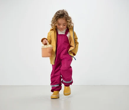 Ein lockiges Kind in gelber Jacke, rosafarbenen Hosen und gelben Gummistiefeln hält eine Gießkanne und steht auf dem Boden.