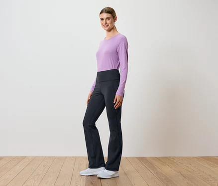 Eine Frau posiert seitlich in einem lila, langärmeligen Oberteil und einer schwarzen Hose mit hohem Bund.
