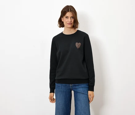Frau steht und trägt einen schwarzen Pullover mit Herzapplikation und Jeans.
