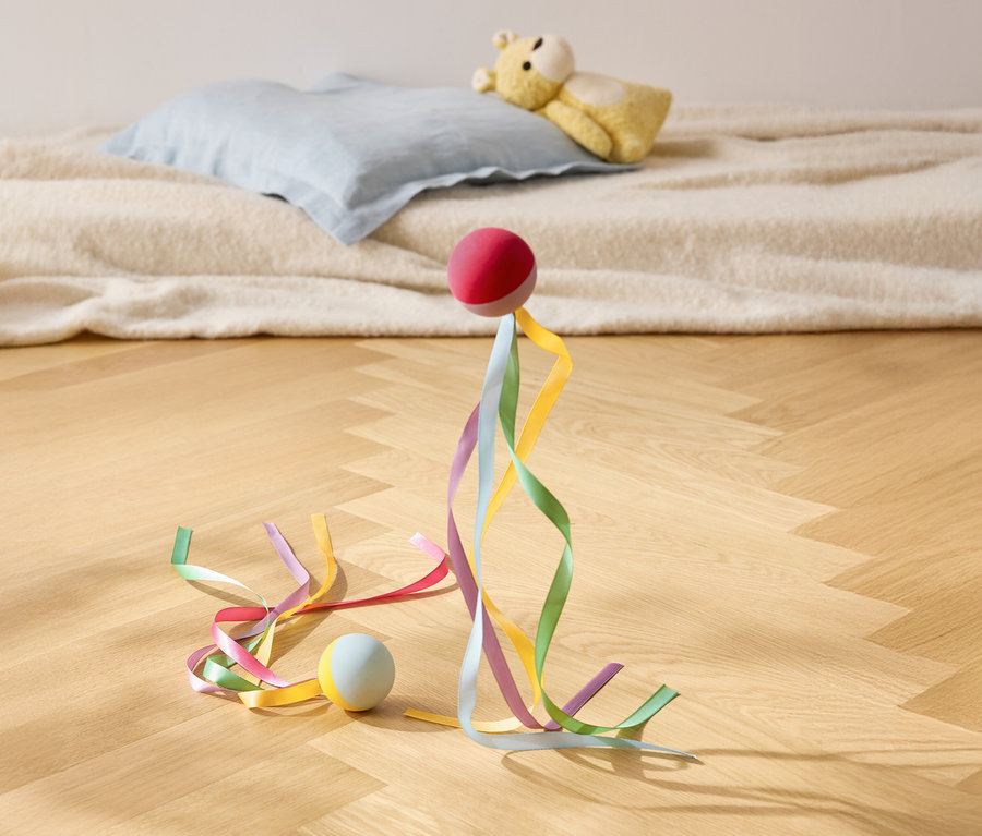Ein roter Ball steht mit bunten Bändern auf einem Holzboden, im Hintergrund ein Kissen und ein Teddybär.