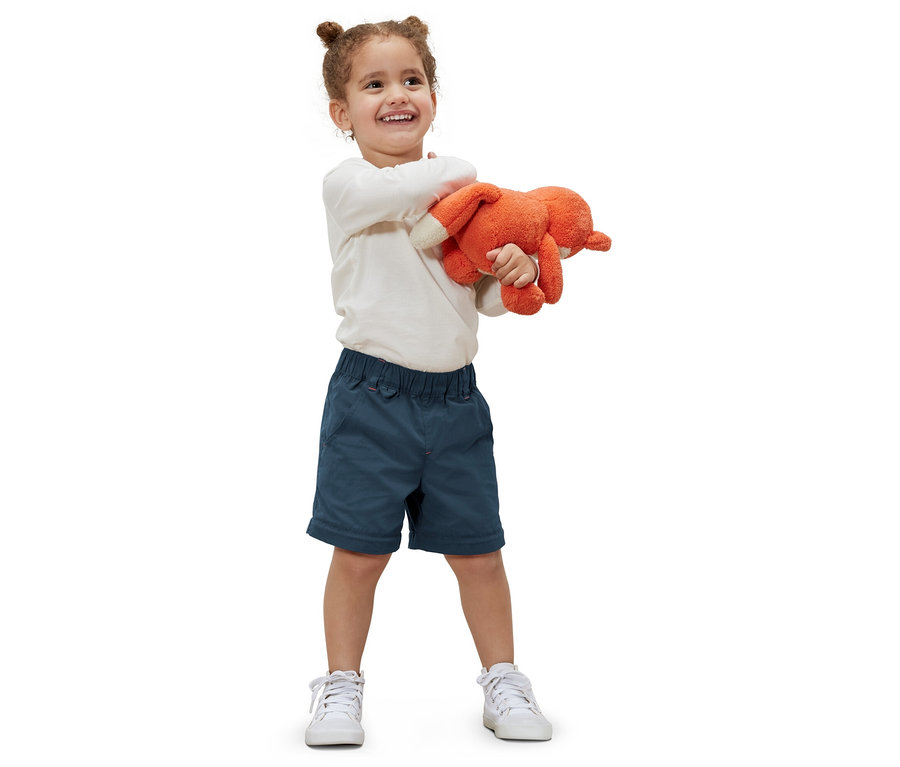 Ein lächelndes kleines Mädchen hält ein orangefarbenes Stofftier und trägt eine Kleinkinder-Funktionshose und weiße Turnschuhe.