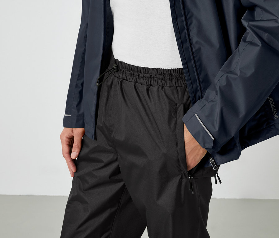 Mann mit Hand in der Tasche einer schwarzen Hose, trägt eine blaue Jacke.