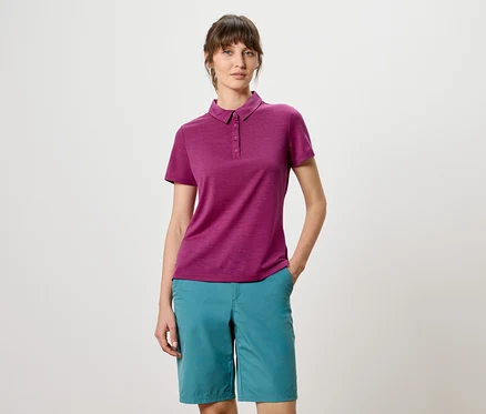 Frau mit weinrotem Poloshirt und türkisfarbenen Shorts posiert vor einem weißen Hintergrund.