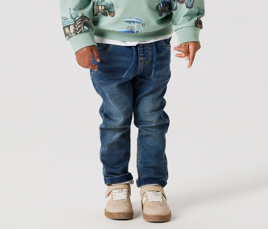 Kind trägt NAME IT Kinder-Sweat-Jeans, hellgrünes Traktor-Sweatshirt und beigefarbene Schuhe vor einem weißen Hintergrund.