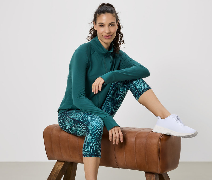 Frau sitzt auf einem Turnbock, trägt grüne Sportjacke, gemusterte Leggings und weiße Turnschuhe.