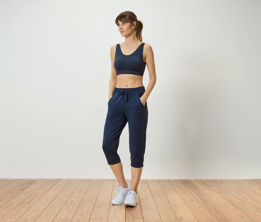 Frau mit marineblauem Sport-BH und marineblauer Hose mit Händen in den Taschen, die weiße Turnschuhe trägt.