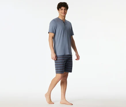 Ein lächelnder Mann trägt ein blaues SCHIESSER Shorty-Set mit Kurzarm-Henley-Oberteil und gestreiften Shorts, barfuß stehend.