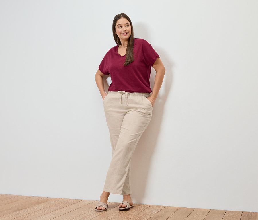 Frau posiert in dunkelrotem Shirt aus Leinenmix und einer cremefarbenen Leinenhose vor einer Wand.