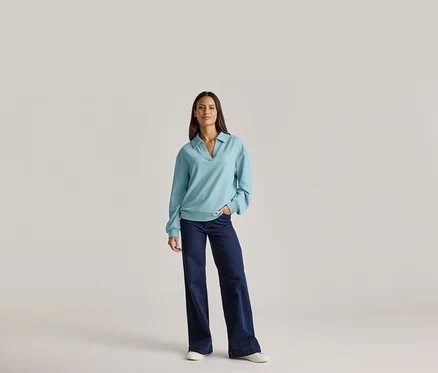 Frau in hellblauem Langarmshirt mit Kragen und dunkler Jeans, eine Hand in der Hosentasche.