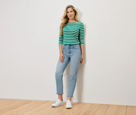 Eine blonde Frau steht vor einer Wand und trägt ein grün-weiß gestreiftes Langarmshirt, hellblaue Jeans und weiße Sneaker.