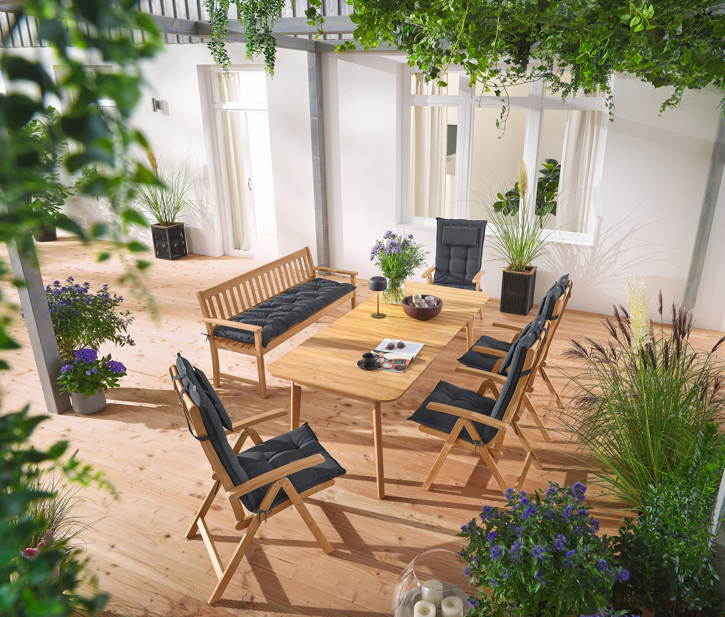 Auf einer Terrasse stehen ein doppelt ausziehbarer Teak-Dining-Tisch, eine klassische Teak-Gartenbank und Teak-Hochlehner mit anthrazitfarbenen Hochlehnerauflagen, sowie ein Pflanztopf mit Textilgeflecht und eine graue Solar-Akku-Tischleuchte.