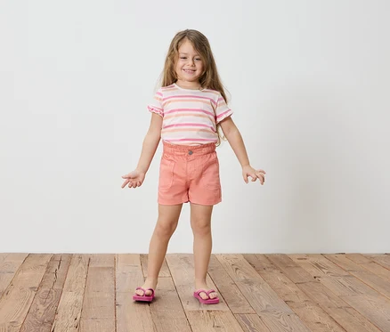 Mädchen mit blonden, langen Haaren, trägt ein gestreiftes T-Shirt, Shorts und Sandalen.