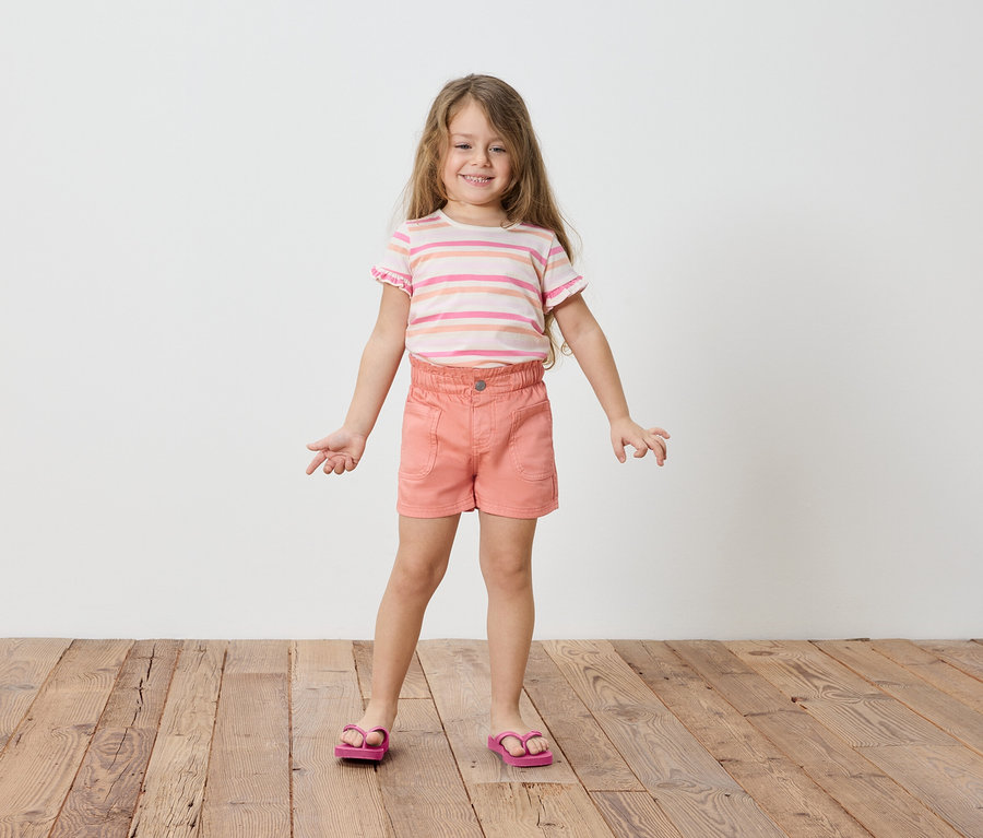 Mädchen mit blonden, langen Haaren, trägt ein gestreiftes T-Shirt, Shorts und Sandalen.