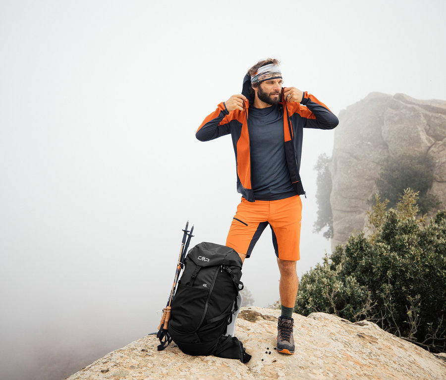 Mann steht mit CMP Trekking-Rucksack HAVRE, 40 Liter auf einem Felsen. Wanderstöcke lehnen am Rucksack.