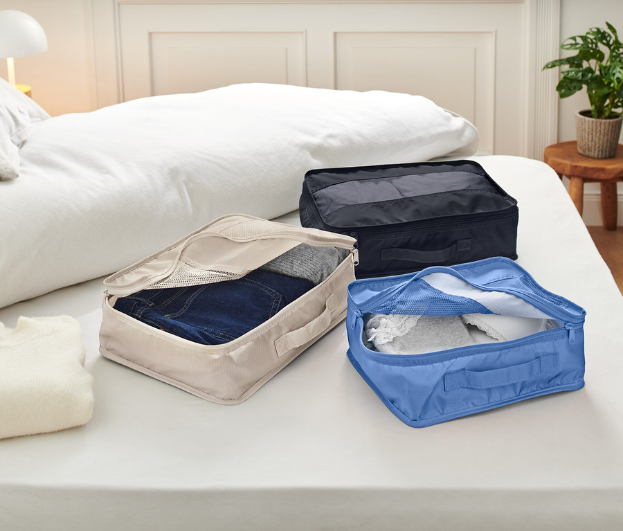 Drei offene Packwürfel liegen auf einem Bett mit weißer Bettwäsche. Die Packwürfel sind beige, blau und schwarz und enthalten Kleidung.