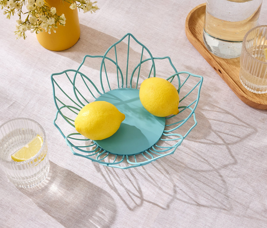 Zwei Zitronen liegen in einer blauen Drahtschale auf einem Tisch neben einem gelben Topf mit Blumen und Gläsern mit Wasser.