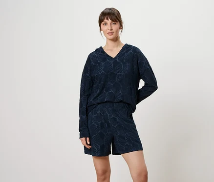 Frau trägt ein marineblaues, gemustertes Langarmshirt mit V-Ausschnitt und Shorts vor einem neutralen Hintergrund.