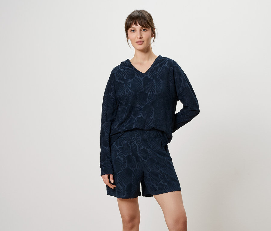 Frau trägt ein marineblaues, gemustertes Langarmshirt mit V-Ausschnitt und Shorts vor einem neutralen Hintergrund.