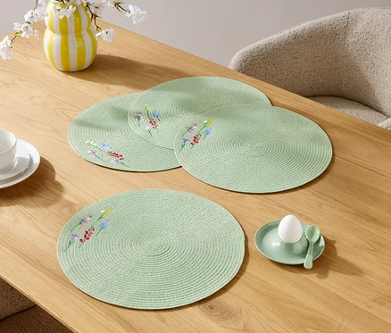 Nahaufnahme von vier runden, hellgrünen Tischsets mit Blumenmuster auf einem Holztisch, zusammen mit einer Tasse, einem Teller, einer Blumenvase und einem Eierbecher mit Ei.