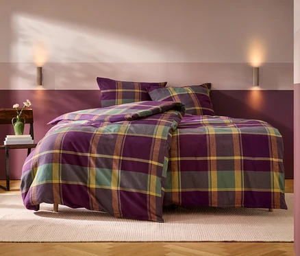 Ein Bett mit lila, grün, gelb und braun karierter Bettwäsche, bestehend aus einem Bettbezug und zwei Kissen. Links daneben steht ein Holzstuhl mit einem Stapel Bücher und einer grünen Vase mit Blumen. Die Wand hinter dem Bett ist zweifarbig gestrichen, unten dunkelviolett und oben hellrosa-violett, mit zwei Wandleuchten. Ein heller Teppich liegt auf dem Holzboden.