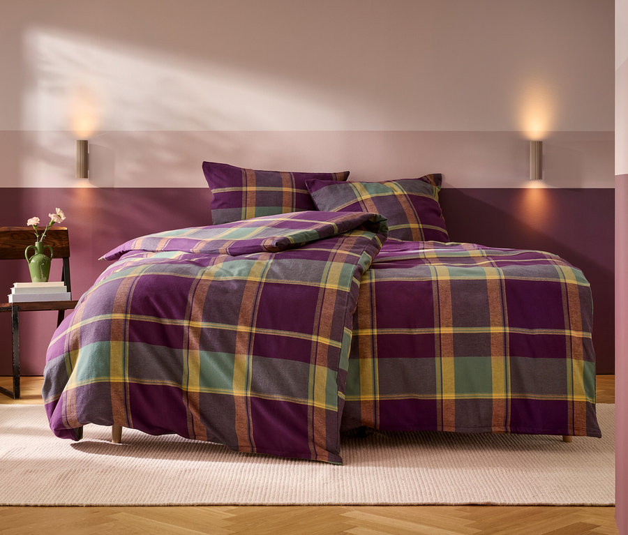 Ein Bett mit lila, grün, gelb und braun karierter Bettwäsche, bestehend aus einem Bettbezug und zwei Kissen. Links daneben steht ein Holzstuhl mit einem Stapel Bücher und einer grünen Vase mit Blumen. Die Wand hinter dem Bett ist zweifarbig gestrichen, unten dunkelviolett und oben hellrosa-violett, mit zwei Wandleuchten. Ein heller Teppich liegt auf dem Holzboden.