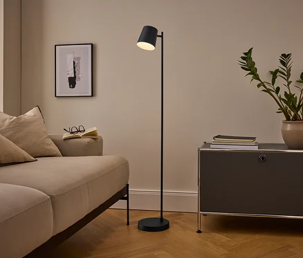 Gemütliches Wohnzimmer mit beigem Sofa, schwarzer Stehlampe und dunkelgrauem Schrank. Ein offenes Buch und eine Brille liegen auf dem Sofa, eine Topfpflanze und Bücher auf dem Schrank. Ein gerahmtes abstraktes Bild hängt an der Wand.