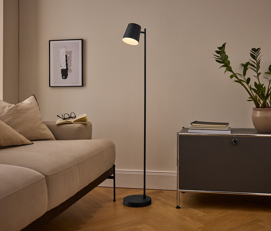 Gemütliches Wohnzimmer mit beigem Sofa, schwarzer Stehlampe und dunkelgrauem Schrank. Ein offenes Buch und eine Brille liegen auf dem Sofa, eine Topfpflanze und Bücher auf dem Schrank. Ein gerahmtes abstraktes Bild hängt an der Wand.