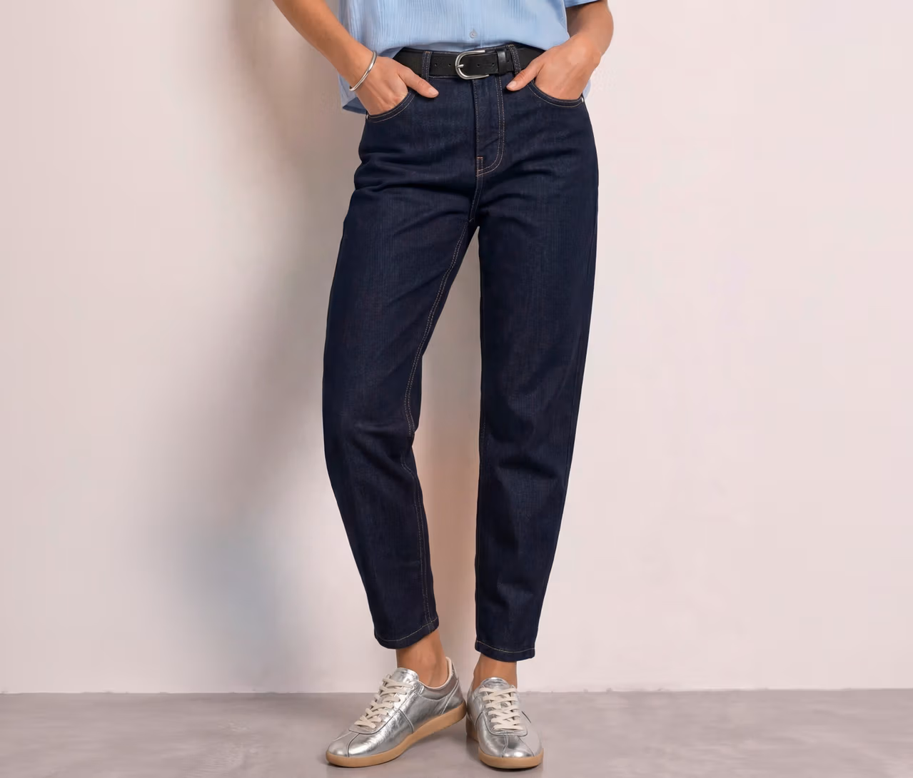 Person trägt dunkelblaue Street One Barrel Jeans, ein hellblaues Oberteil, einen schwarzen Gürtel und silberne Sneaker, die Hände in den Taschen.