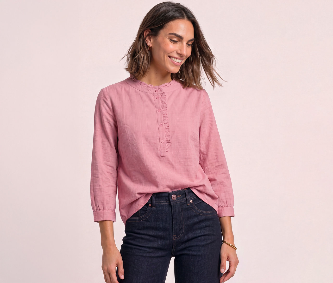 Eine lächelnde Frau trägt eine rosafarbene Street One Cordbluse mit Rüschen und dunkle Jeans.