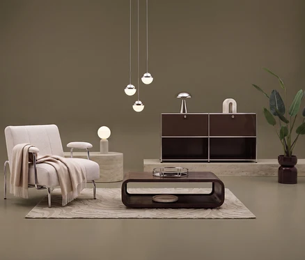 Ein beiger Sessel mit Armlehne und einer cremefarbenen Wohndecke »PLENTY« steht auf einem cremefarbenen Teppich »Cosmo «Waves. Davor ein brauner Couchtisch. Im Hintergrund ein braunes Sideboard Metall »CN3« mit 2 Klappenfächern und 2 offenen Fächern, darauf eine Tischleuchte »Bovista«. Drei Hängelampen und eine Tischleuchte »Traventa« beleuchten den Raum.