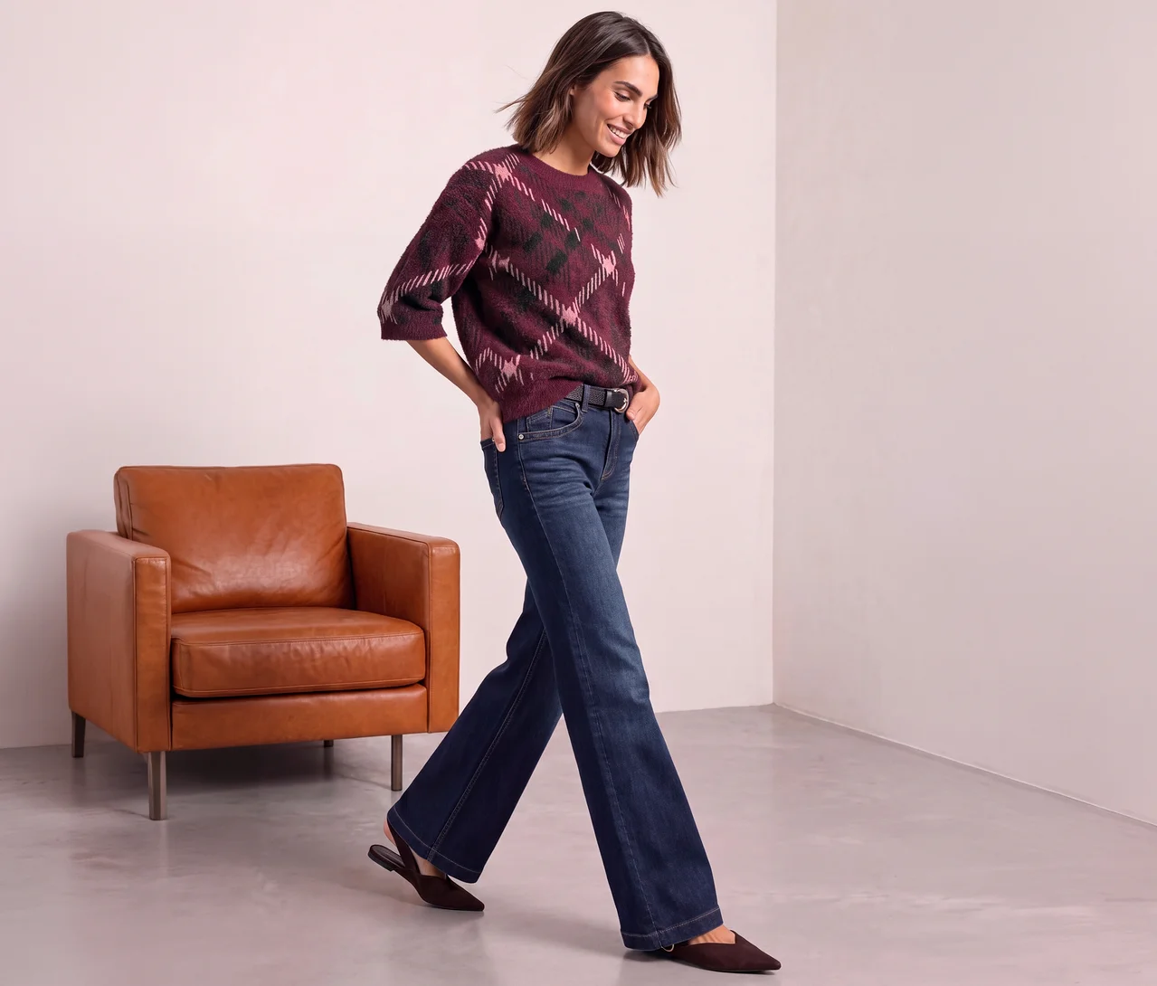 Frau trägt einen Street One Strickpullover mit Argyle-Muster in Bordeaux, Pink und Dunkelgrün, dunkle Jeans und braune Flats, geht an einem braunen Ledersessel vorbei.