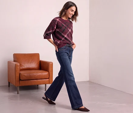 Frau trägt einen Street One Strickpullover mit Argyle-Muster in Bordeaux, Pink und Dunkelgrün, dunkle Jeans und braune Flats, geht an einem braunen Ledersessel vorbei.
